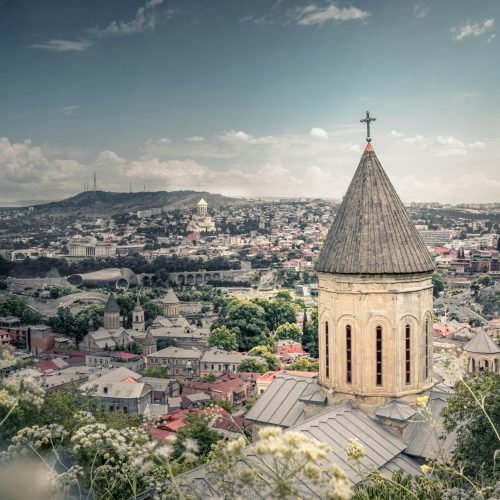 الرئيسية View of a historical church tower with Tbilisi's cityscape in the background under a blue sky.